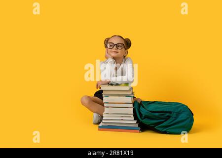 Ragazza della scuola elementare cute seduta alla pila dei libri sopra lo sfondo di Yellow Foto Stock