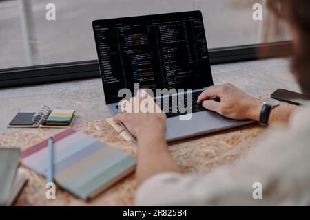 Primo piano delle mani dei programmatori maschi scrivono il codice sul laptop seduto in un bar vicino alla finestra Foto Stock