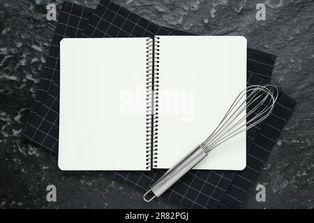 Libro di ricette in bianco e frusta su un tavolo con texture nere, piatto. Spazio per il testo Foto Stock