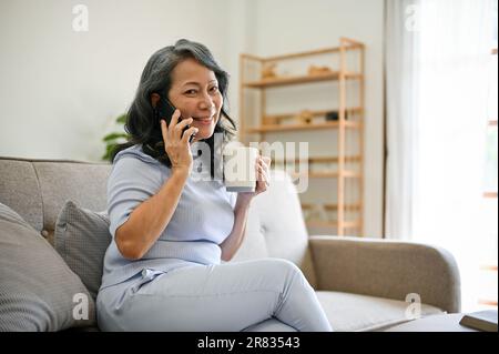 Una donna asiatica di mezza età attraente e allegra ama il caffè mattutino e parlare al telefono con la sua amica mentre si rilassa su un divano in lei Foto Stock