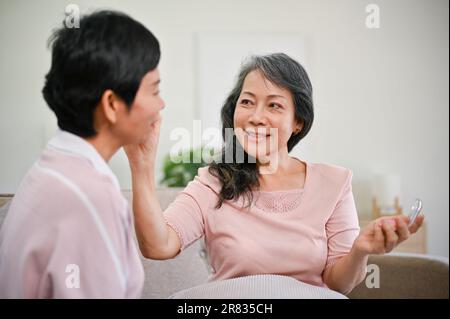 Una donna asiatica di mezza età attraente e felice che applica il trucco alla sua amica, aiutando la sua amica a fare il trucco, e trascorrere il tempo divertente a casa la settimana Foto Stock