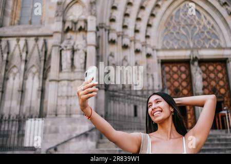 Donna latino-americana ottimista che tocca la testa e sorride mentre si prende selfie via smartphone contro l'ingresso della cattedrale medievale di giorno a Barcellona, Spagna Foto Stock