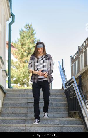 Giovane uomo dai capelli lunghi in camicia a quadri e jeans scende le scale Foto Stock