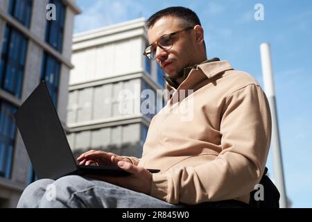Vista dal basso angolo del giovane con disabilità che si concentra sul suo lavoro online su un notebook all'aperto Foto Stock