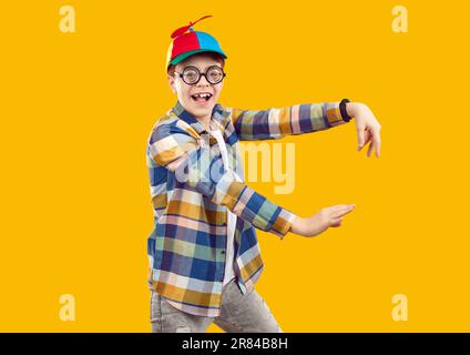 Felice bambino divertente in camicia, cappello di elica e bicchieri che ballano su sfondo arancione Foto Stock