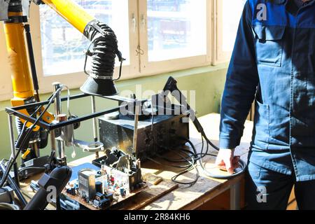 Un elettricista che lavora in veste lavora professionalmente su un tavolo per la riparazione in un'officina per la rimozione di componenti elettronici e la produzione di parti e spa Foto Stock