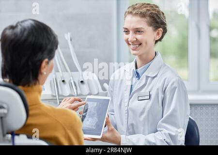 Ritratto di un dentista sorridente che mostra immagini radiografiche su tablet alla paziente in clinica Foto Stock