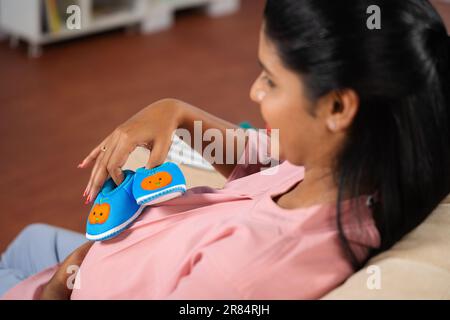 Shoulder shot di donna incinta indiana che gioca con le scarpe del bambino o Booties sul pancino a casa - concetto di connessione prenatale, maternità e gravidanza Foto Stock