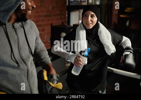 Felice giovane donna musulmana con una bottiglia d'acqua guardando afro-americana maschio boxe allenatore in grigio durante la pausa Foto Stock