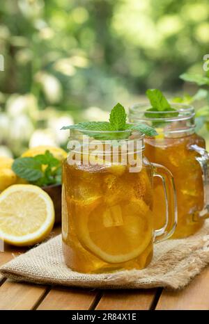 Tè ghiacciato in vasetti di vetro con fettine di limone e menta all'aperto su un tavolo di legno con sfondo naturale Foto Stock