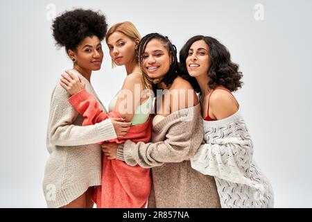 Ritratto di donne multietniche eleganti e fiduciose in maglioni a maglia sorridente e abbracciante isolato sul grigio, diversi tipi di corpo e auto-accettazione Foto Stock