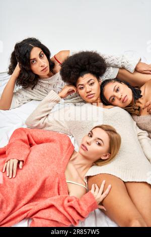 Vista ad alto angolo delle donne multietniche in comodi jumper a maglia che guardano la telecamera mentre sdraiati sul letto isolato sul grigio, diversi tipi di corpo e auto-acce Foto Stock