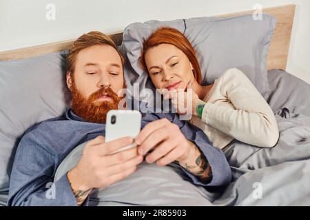 coppia rossa che trascorre il tempo senza bambini, giorno libero, marito e moglie, uomo bearded che usa lo smartphone vicino alla donna, vita confortevole, camera da letto accogliente, caref Foto Stock