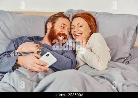 spensierato, risate, tempo dello schermo, coppia tatuata che trascorre il tempo senza bambini, giorno libero, marito e moglie, uomo bearded e donna redhead, utente dello smartphone Foto Stock