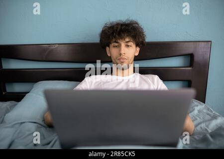 Ritratto di un giovane arabo seduto a letto con un computer portatile, un ragazzo che utilizza il computer portatile in camera da letto per il lavoro online e godere di freelancing Foto Stock