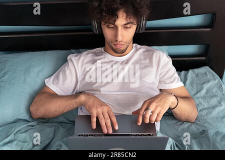 Felice giovane uomo che guarda le serie sul PC seduto a letto a casa, godendo di contenuti o musica in cuffie, studiando online, lavoro freelance Foto Stock