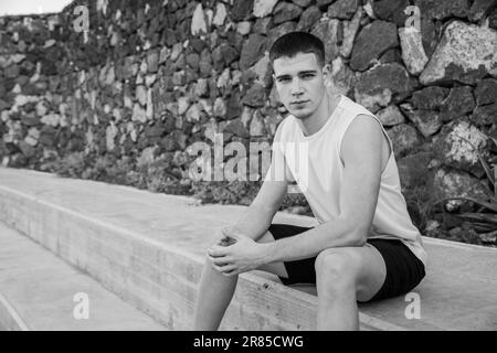 Un ragazzo adolescente è seduto al bordo di un campo di pallacanestro, foto in bianco e nero Foto Stock