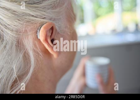 Primo piano di una donna anziana che indossa un dispositivo per l'udito dietro l'orecchio o un dispositivo per la preparazione di bevande calde a casa Foto Stock