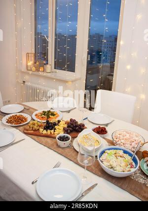 Tavolo festivo decorato con tovaglia bianca e dorata, con assortimento di insalate, vassoio di formaggi, salmone affumicato, bicchieri di vino, bokeh l Foto Stock