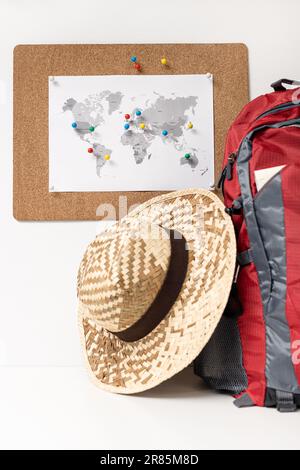 Pianificazione del concetto di destinazione di viaggio. Corkboard con mappa del mondo e spille sulla parete, zaino e cappello estivo Foto Stock