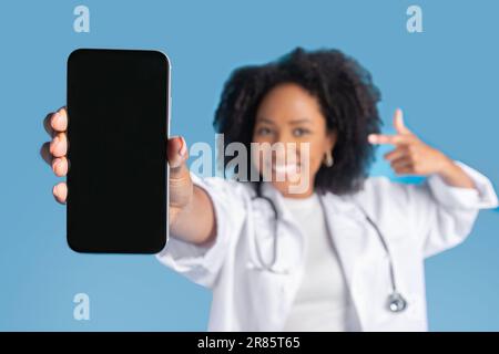 Felice eccitato giovane schiena curly signora medico in cappotto punto dito al telefono, concentrarsi sullo schermo vuoto Foto Stock