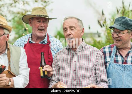 Un vivace gruppo di amici anziani, tutti uomini caucasici, si riuniscono all'aperto per un'escursione piena di divertimento. Cantano insieme con gioia mentre si godono il loro rinfresco Foto Stock