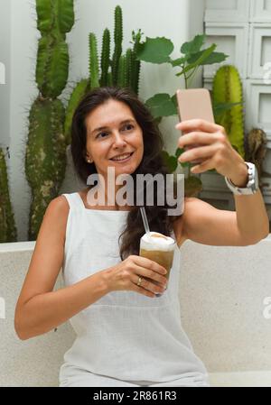 Ritratto di una donna russa di 39 anni senza trucco seduto con un bicchiere di caffè ghiacciato in abito bianco e scattando un selfie Foto Stock