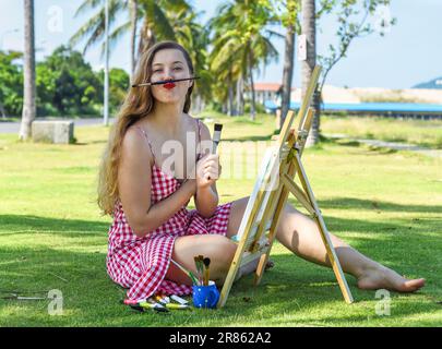 Giovane artista donna che fa un volto divertente con matita e cavalletto vicino al mare e alle palme in estate Foto Stock