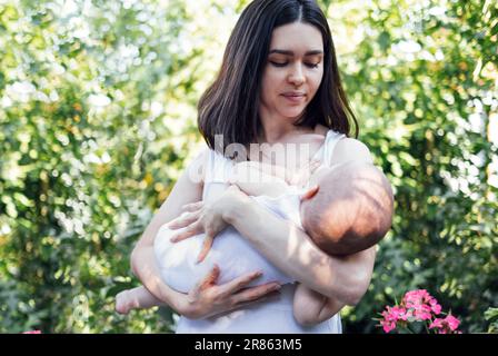 Una bella madre in abiti bianchi sta oscillando il suo bambino tra le braccia in giardino. Un'affascinante bruna donna tiene in braccio un bambino piccolo. A y Foto Stock