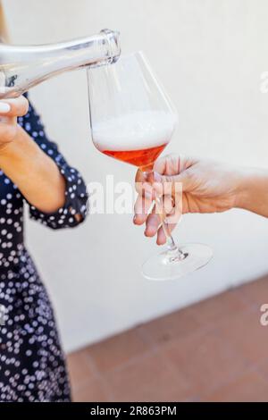 Una giovane donna versa champagne rosa da una bottiglia trasparente. Primo piano di una mano femminile con una manicure ordinata che tiene un elegante bicchiere con un gambo lungo Foto Stock