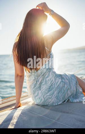 Una giovane donna in una bellissima ed elegante sundress siede sul ponte di una nave e incontra l'alba. Una sottile brunetta gode il mare e il viaggio sul Foto Stock