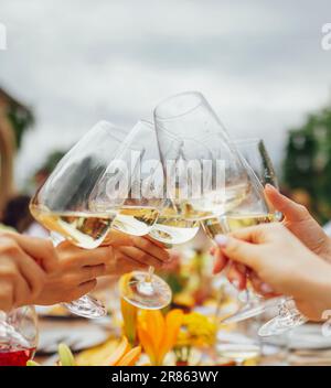 Le mani femminili tengono eleganti bicchieri con gambo lungo di champagne e li stringono sullo sfondo di un tavolo con spuntini e fiori. Fare un cel Foto Stock