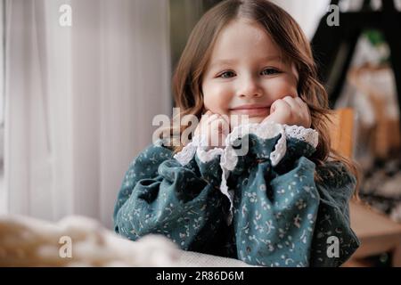 Adorabile bambina con capelli ricci indossa abiti festosi stile rurale è sorridente, guardando la macchina fotografica, seduto accanto al tavolo e sostiene il mento con ha Foto Stock