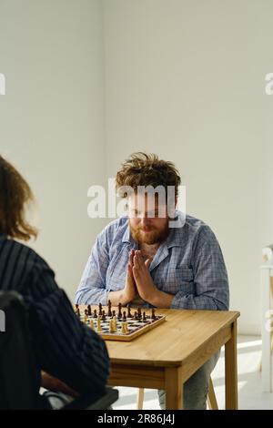 Giovane uomo bearded tenendo le mani messe insieme dal petto e guardando la scacchiera mentre pensa al movimento seguente durante il gioco con l'altro paziente Foto Stock