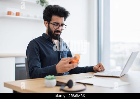 Bella persona matura con occhiali che regge una bottiglia di pillole mentre è seduto in ufficio domestico con un computer portatile a mezzogiorno. Uomo d'affari indiano che esamina la medicina prescritta dopo la consultazione on-line dal medico. Foto Stock