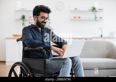 Gioioso uomo indiano in sedia a rotelle che tiene il computer portatile in ginocchio mentre si trova nel mezzo di una luminosa cucina open space. Felice giovane adulto che naviga in Internet pagine durante il recupero dopo un incidente a casa. Foto Stock