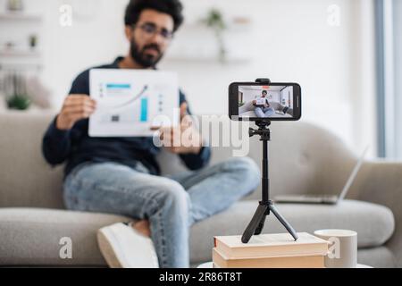 Bel ragazzo indiano in abiti informali seduto sul divano con un report finanziario grafico in mano mentre registra video su smartphone moderno. Finanziatore di successo che parla di statistiche sulla fotocamera. Foto Stock