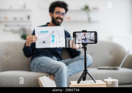 Bel ragazzo indiano in abiti informali seduto sul divano con un report finanziario grafico in mano mentre registra video su smartphone moderno. Finanziatore di successo che parla di statistiche sulla fotocamera. Foto Stock