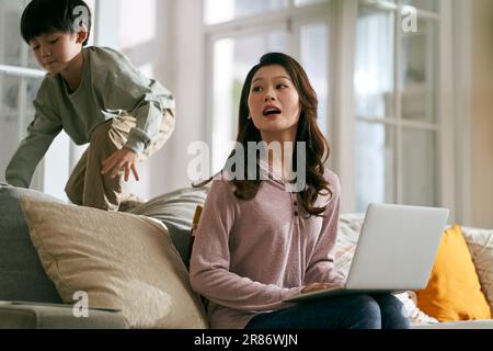 giovane madre asiatica d'affari cercando di lavorare a casa infastidita da figlio iperattivo Foto Stock