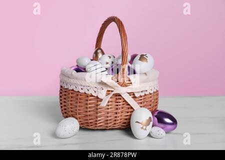 Cestino di vimini con uova di Pasqua decorate in modo festivo su un tavolo di marmo bianco su sfondo rosa Foto Stock