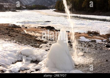 Un getto d'acqua scorre giù su un alto blocco di ghiaccio in piedi sulla riva di un fiume ghiacciato che scorre attraverso una valle di montagna in una serata d'inverno. Katun Foto Stock