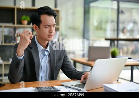 Un pensieroso uomo d'affari asiatico millenario si concentra sulle sue attività sul suo notebook, guardando il suo notebook con un volto serio e lavorando alla sua scrivania Foto Stock