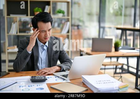 Un uomo d'affari asiatico, stressato e premuroso, sta guardando lo schermo del suo computer portatile con un volto serio, facendo un piano per risolvere il problema Foto Stock