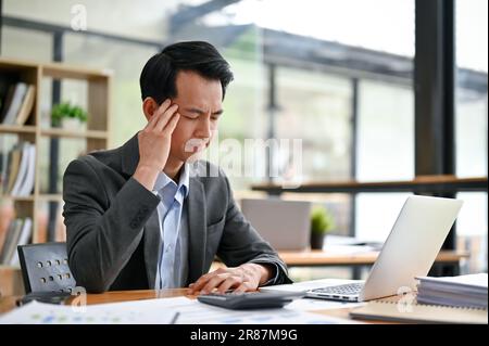 Un uomo d'affari asiatico, un millennio stressato e premuroso, sta facendo un piano per risolvere i problemi, lavorando alla sua scrivania in ufficio. Foto Stock