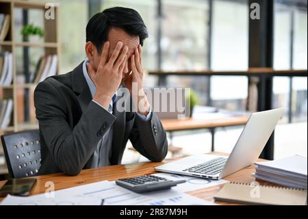 Un uomo d'affari asiatico, stressato e frustrato, sta coprendo il suo volto, facendo un piano penosamente per risolvere i problemi, sconvolto dal suo progetto fai Foto Stock