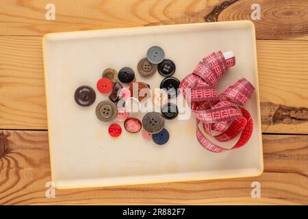 Molti bottoni in un contenitore di plastica e metro a nastro per i marinai su un tavolo di legno, macro, vista dall'alto. Foto Stock