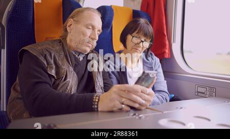 La coppia anziana viaggia in treno, l'anziano tiene in mano un telefono cellulare, entrambi guardano lo smartphone e parlano l'uno con l'altro Foto Stock