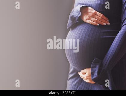 il primo piano della pancia di una donna incinta. Le mani delle ragazze abbracciano il ventre su sfondo blu. Concetto di salute materna. Foto Stock