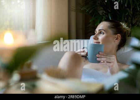 donna che si rilassa nel bagno e beve un caffè nel bagno di casa. guardando fuori dalla finestra Foto Stock