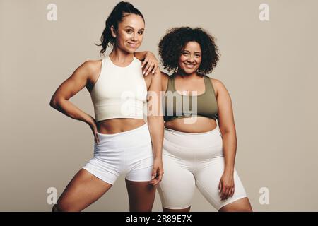 Due giovani donne in forma con corpi diversi si incontrano nello studio, indossando abbigliamento sportivo. Giovani femmine che mostrano con orgoglio la loro dedizione ad un sano Foto Stock